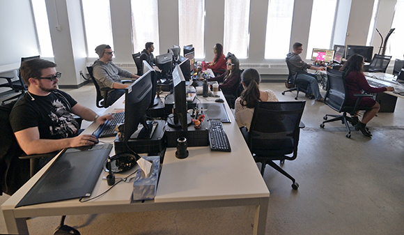 Several people sit at long white desks using computers in a bright office with large windows.