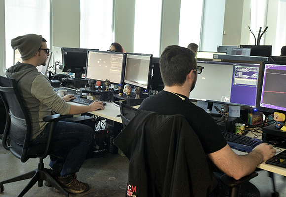 Two men sit at desks with multiple computer monitors in a bright office space.