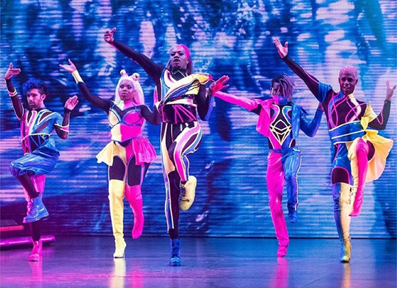 Five dancers in vibrant, patterned neon costumes perform a choreographed routine on a lit stage.