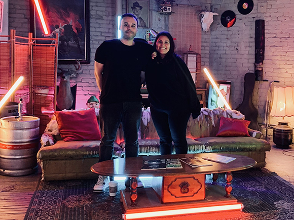 A man in a black t-shirt and a woman in black clothing stand in a room with neon lights and vintage decor.