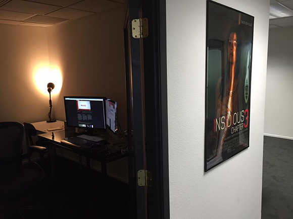 A view into a dark office containing a desk, computer monitors, and a lamp, seen through an open doorway.
