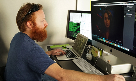 A man with a red beard sits at a desk using a laptop and two large computer monitors.