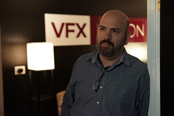 A bald man with a beard and a blue button-down shirt stands in front of a sign that reads VFX.