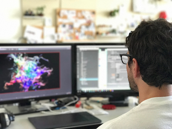 A man with dark hair and glasses looks at two computer monitors displaying colorful graphics and software.