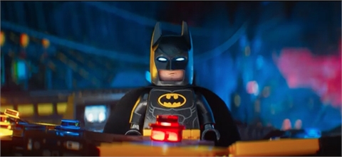 A Lego Batman minifigure stands in front of a blurred, colorful city background.