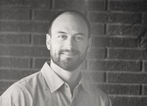 Black and white portrait of a smiling man with a beard wearing a collared shirt against a brick wall.