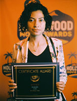 A woman with dark curly hair holds a framed certificate award against an orange Hollywood Movie Awards backdrop.