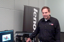 A man in a black shirt gestures toward an open computer tower next to a Lenovo banner.