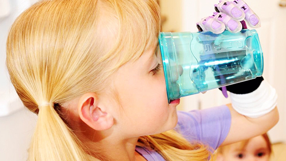 A blonde girl in a purple shirt drinks from a translucent blue cup held by a hand in a purple and white glove.