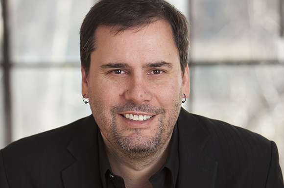 A smiling man with dark hair and a light beard wearing a black blazer and small hoop earrings.