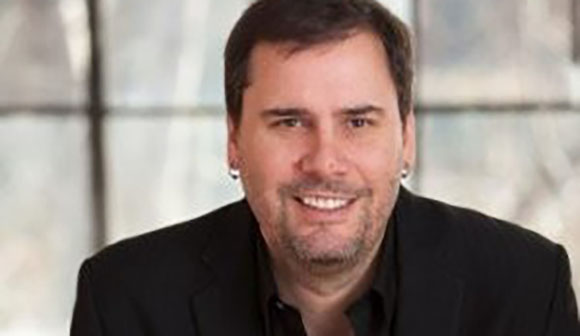 A man with short dark hair and a light beard smiling while wearing a black blazer.