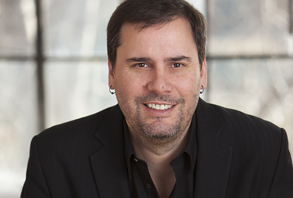 A man with short dark hair and a light beard wearing a black blazer, smiling at the camera.