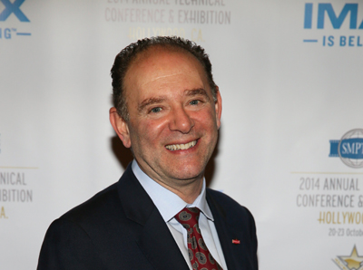 A smiling man in a dark suit, light blue shirt, and patterned red tie stands before a white backdrop.