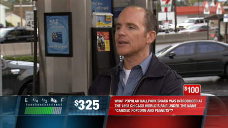 A man in a blue jacket speaks in front of a drive-thru window with a trivia question graphic at the bottom.