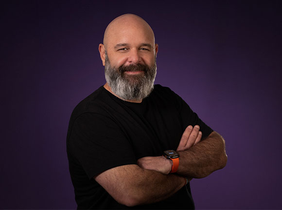 A bald man with a grey beard and a black t-shirt smiling against a purple background.