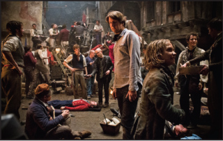 Actors and crew members on a dimly lit, historical-style film set with various props and equipment.