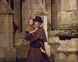A man in a brown robe and top hat carries a young girl in a tan cloak through stone ruins.