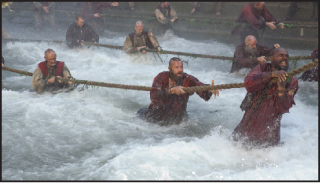 Men in period clothing struggle through turbulent white water while gripping thick ropes.