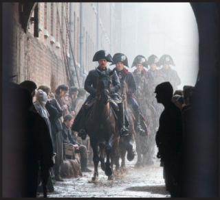 Soldiers in historical military uniforms ride horses down a narrow, misty street past onlookers.