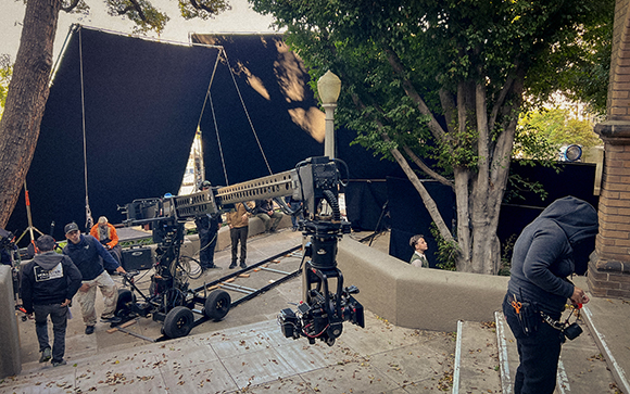 Film crew members work around a camera crane and large black backdrops on an outdoor set.