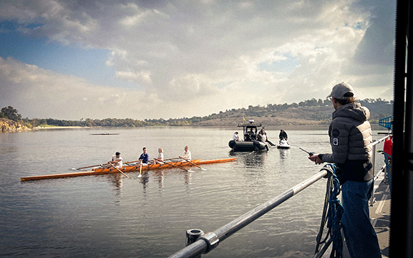 A rowing crew in a long shell on a calm lake under a cloudy sky.