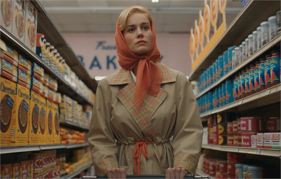A woman wearing an orange headscarf and tan trench coat stands in a grocery store aisle.