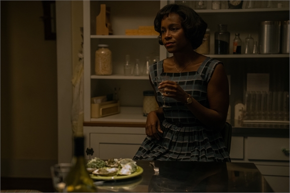 A woman in a blue plaid dress sits at a table holding a glass of wine in a dimly lit kitchen.
