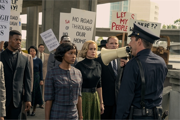 A group of diverse protesters hold signs and face a police officer holding a megaphone.