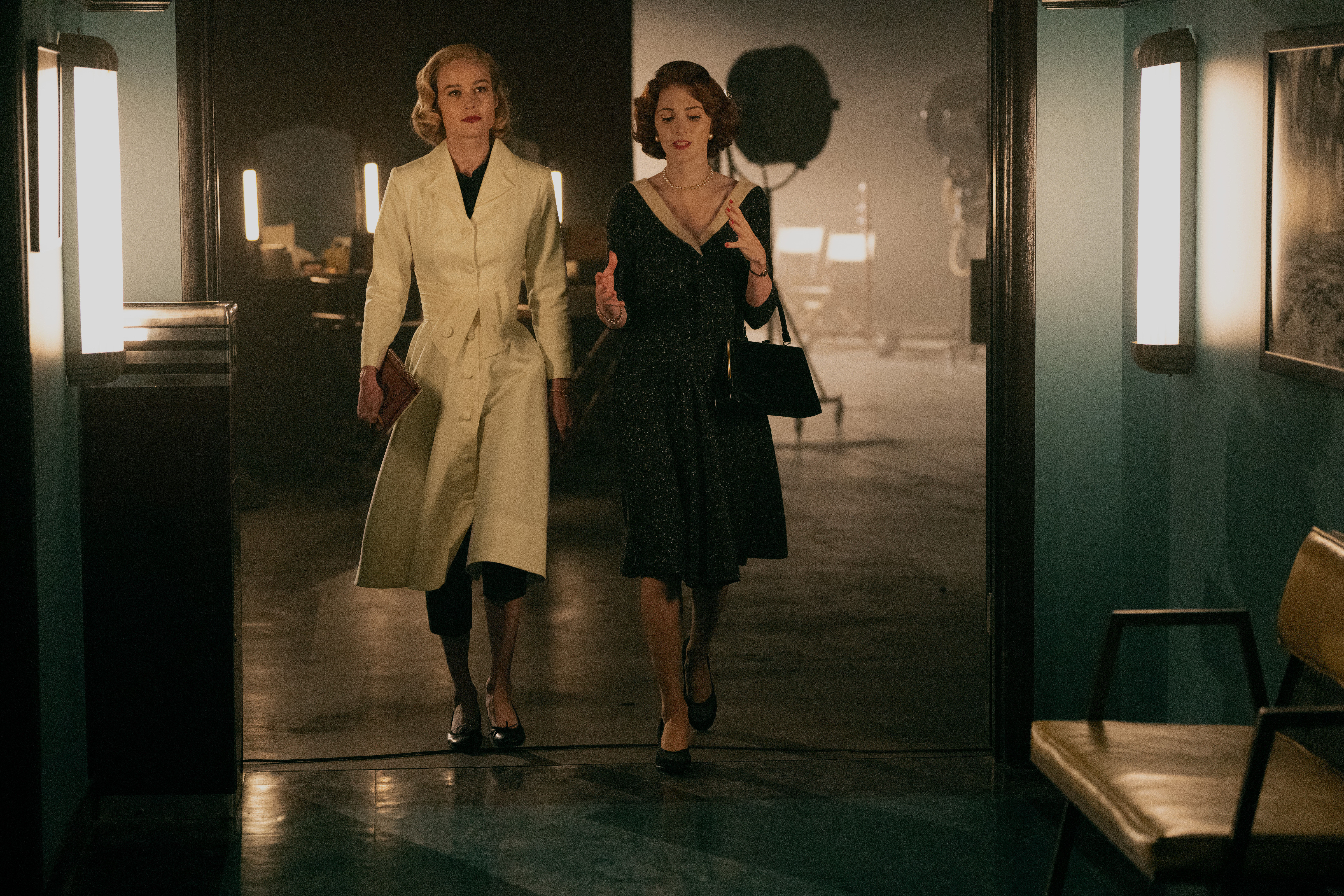 Two women in vintage attire walk through a doorway toward the camera in a dimly lit studio.