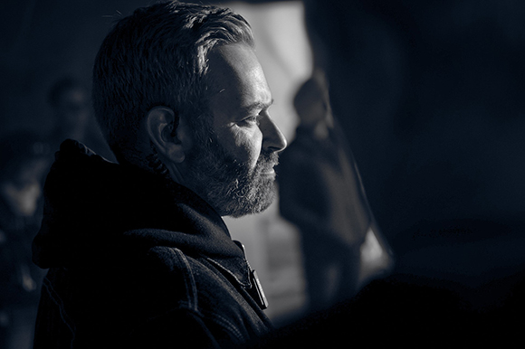 Side profile of a middle-aged man with a beard looking toward a light source in a dark setting.