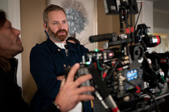 A bearded man in a dark jacket looks toward a professional film camera being operated by someone in the foreground.