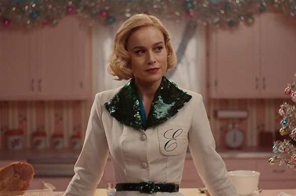 A blonde woman in a white jacket with a large green sequined collar stands in a pink kitchen.