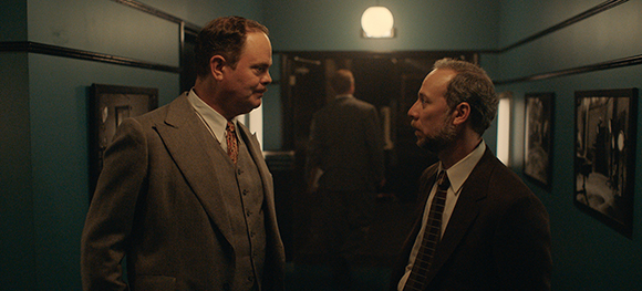 Two men in vintage suits talk in a dimly lit teal hallway with framed pictures on the walls.