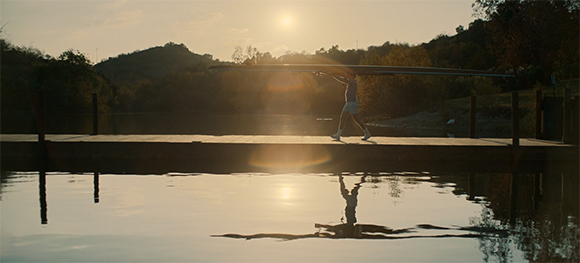 Silhouette of a person carrying a long boat across a wooden dock toward a calm lake at sunset.