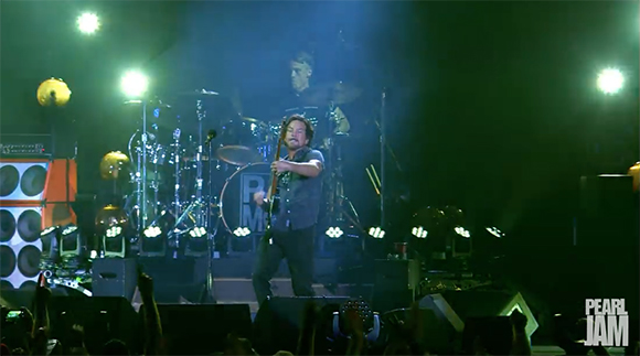 A man plays an electric guitar on a dark stage illuminated by bright spotlights and stage fog.