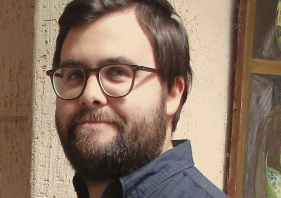 A man with dark hair, glasses, and a full beard smiling slightly.