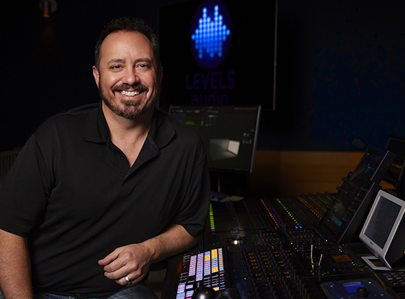 A smiling man in a black polo shirt sits in a dark studio next to a large audio mixing console.