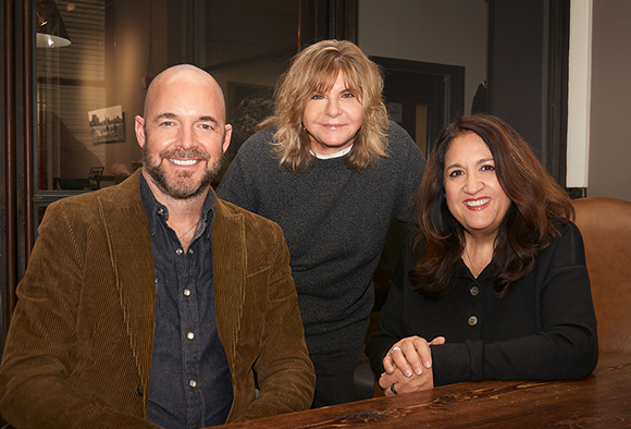 A man in a corduroy jacket and two women smiling together in an indoor setting.