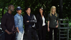 Five people stand in a row outdoors in front of dense green foliage.