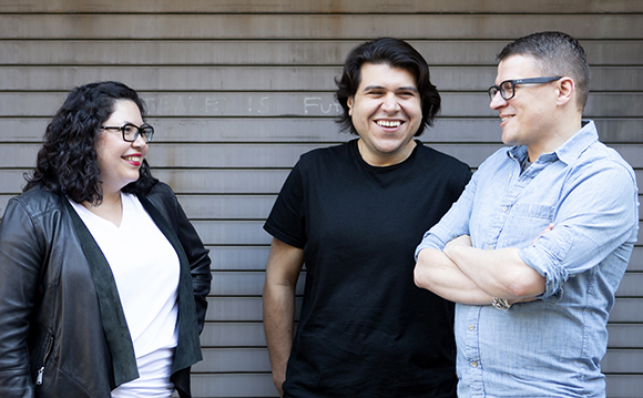 Three smiling people stand together in front of a corrugated metal wall.