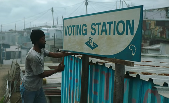 A man in a grey shirt and black beanie stands beside a large sign that reads VOTING STATION.