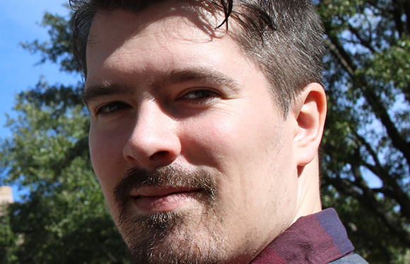 Close-up portrait of a man with dark hair, brown eyes, and a goatee against a background of green trees.