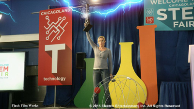 A woman stands before a blue curtain with large STEM fair graphics and laboratory glassware cutouts.
