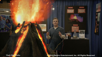 A person stands beside a model volcano erupting with bright orange and yellow light in a science fair setting.
