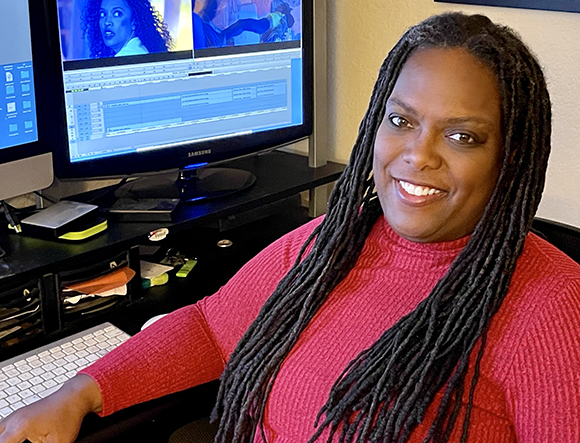 A smiling woman with long dreadlocks wearing a red sweater sits at a desk with dual computer monitors.