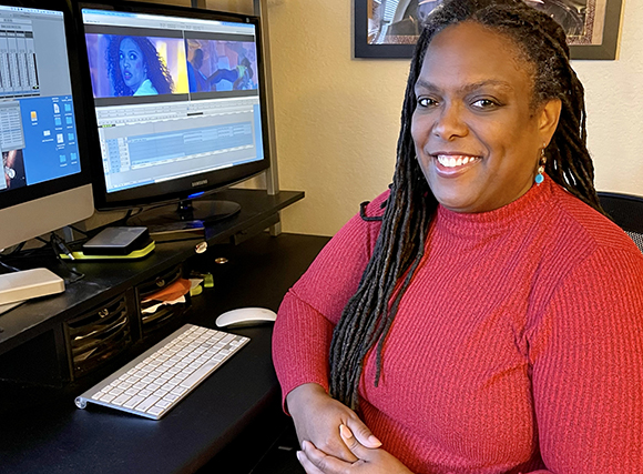 A smiling woman with long dreadlocks wearing a red shirt sits at a desk with two computer monitors.