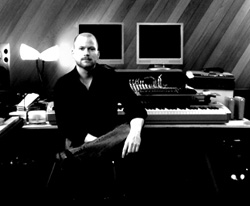 A man in a black shirt sits in a music studio with monitors and a keyboard.