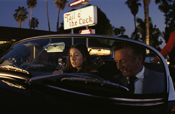 A man and woman sit in a dark convertible at night under a glowing Tail o' the Lock sign.