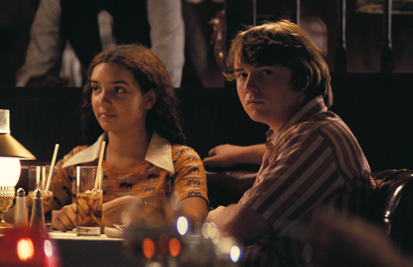 A young woman and a young man sit at a restaurant table with drinks in a dark setting.