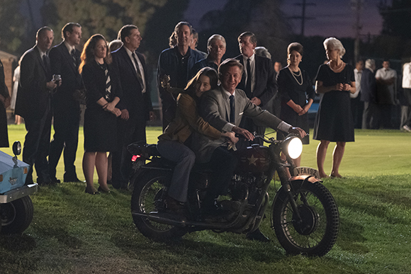 A man and woman sit on a vintage motorcycle in a field at night, surrounded by a crowd of people.